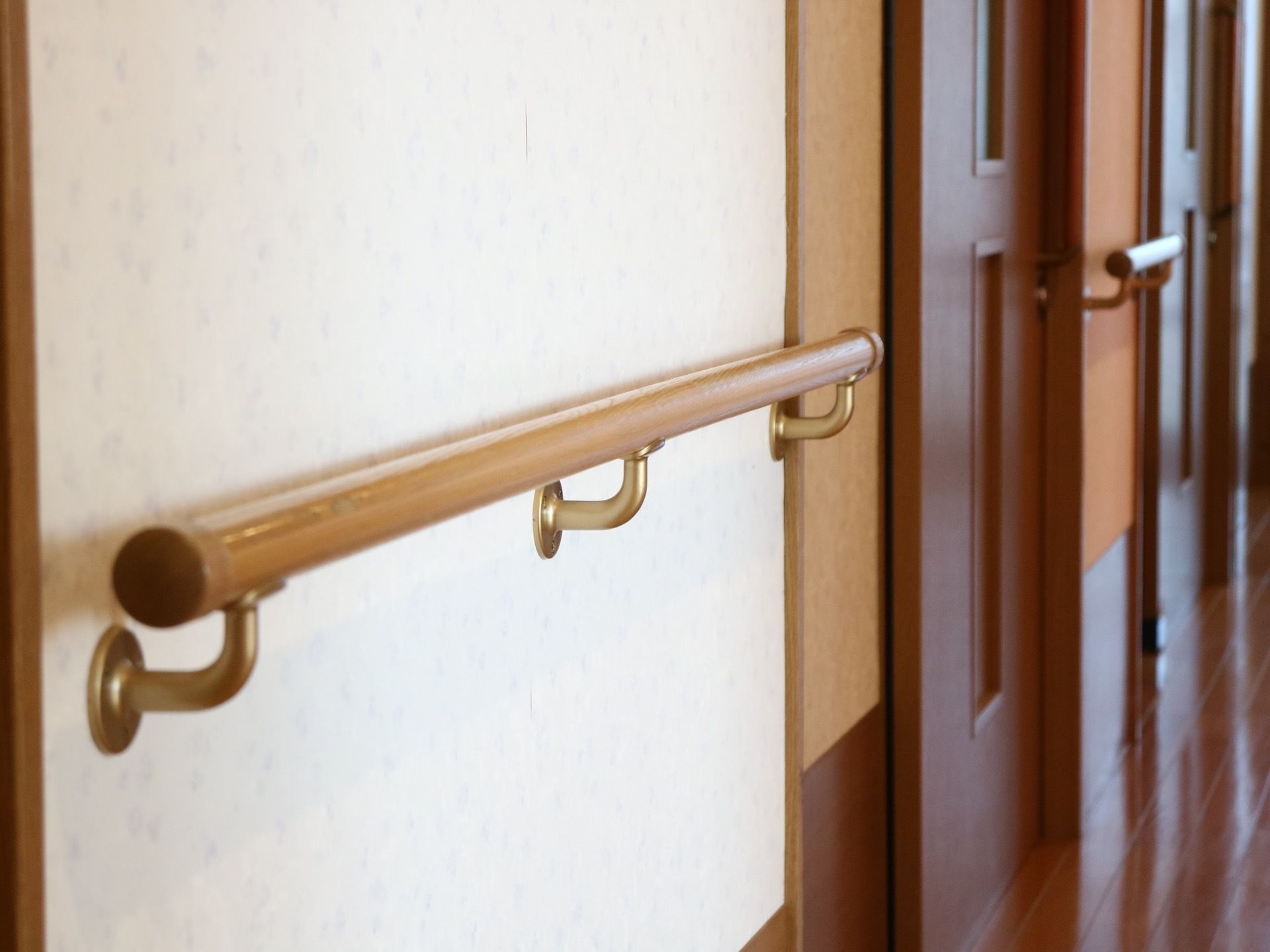 廊下の壁は白い壁紙が貼られている。奥に木製の引き戸が見えている。ドアの間の壁に手すりが取り付けられている。