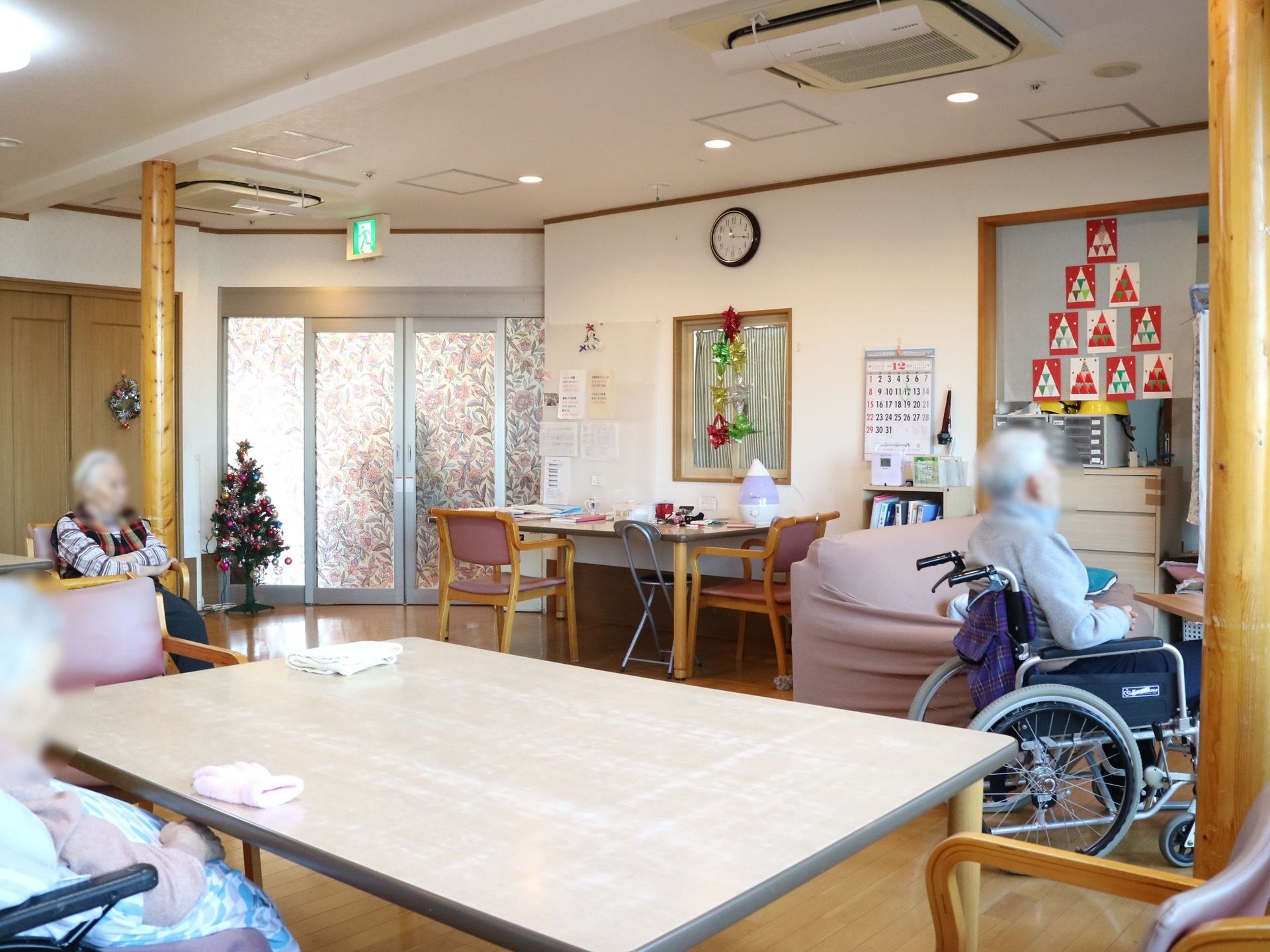 左奥に入口があり、天井に非常口の表示がされている。手前に大きな四角いテーブルがあり、入居者様が椅子に座っている。