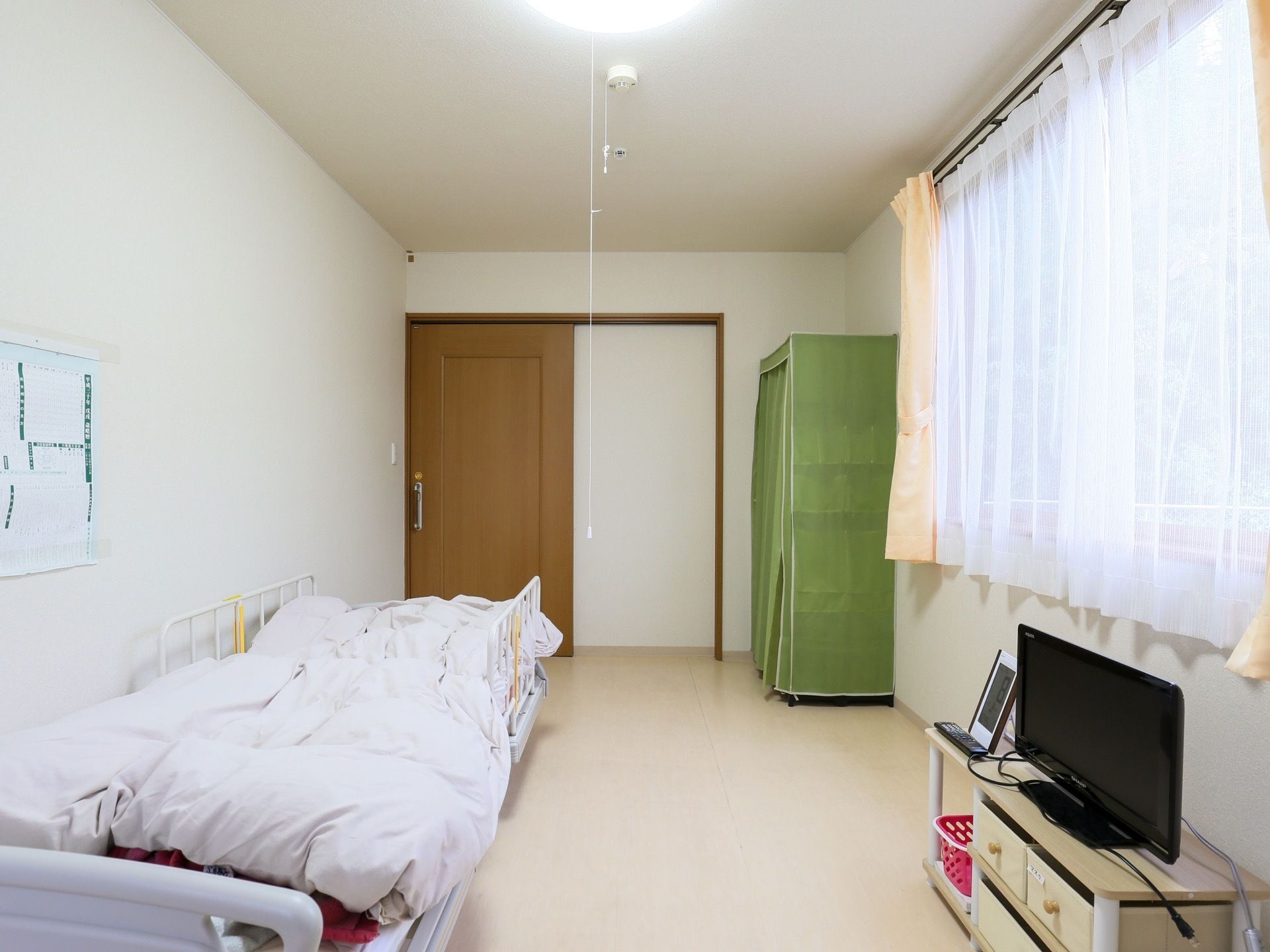 居室の一例である。右側に大きな窓があり、レースのカーテンが閉められている。左手の壁際に介護用ベッドがある。
