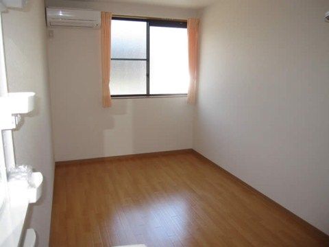 明るい色のフローリングの床、白い壁の居室である。エアコンが設置され、窓にはオレンジ色のカーテンが掛けられている。