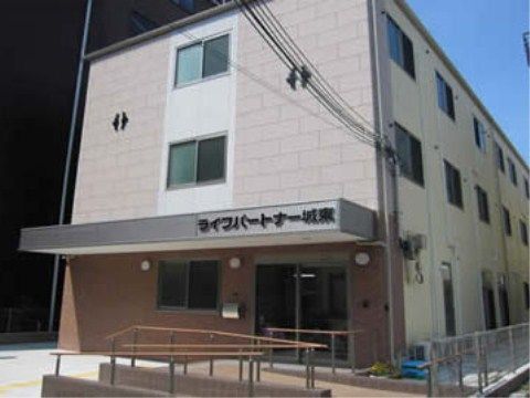 地上3階建て、箱型でピンクベージュ色の外壁の建物の玄関側