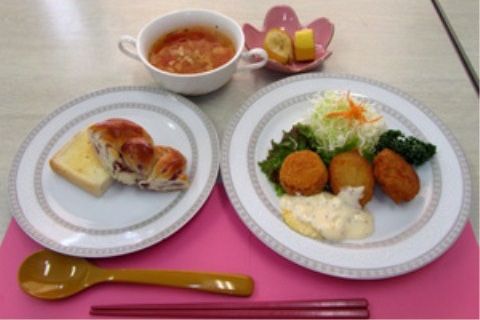 洋風の献立である。お皿にパン、揚げ物、果物が盛られ、カップにはスープが入っている。箸とスプーンが用意されている。