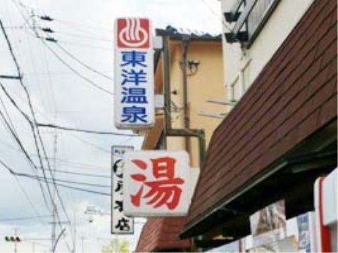 歩道の上に、温泉施設の看板が出ている。手前には酒屋、奥には飲食店がある。施設は周囲に高い建物のない住宅街に位置している。