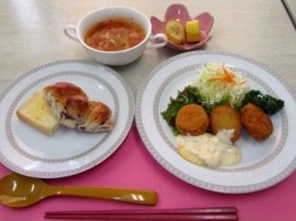 パン食の洋風の献立である。お皿にパンや揚げ物、果物が盛られ、カップにはスープが入っている。箸とスプーンが用意されている。