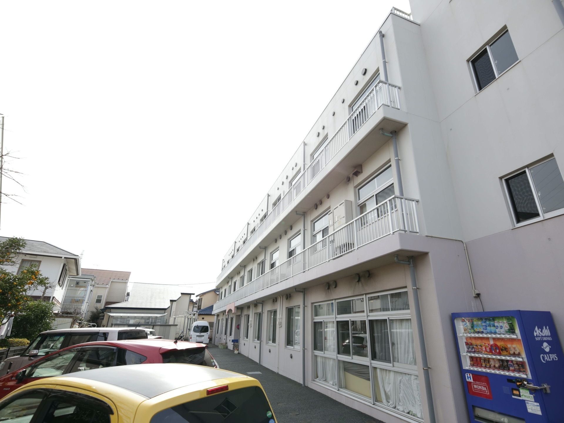 白い外壁でシンプルなデザインの施設は3階建てで、各居室にはベランダが設置してある。駐車場には多くの車が停まっている。