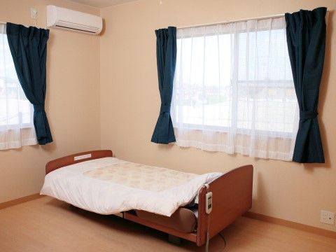 右と左に窓があり、レースカーテンが引かれている。部屋の角にベッドが設置されている。ベッドにはリモコンが吊るされている。