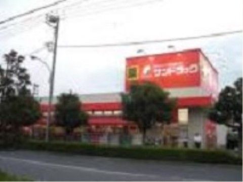 地域のドラッグストアの店舗の外観である。平屋建ての建物で、屋上には大きな看板が設置されている。周囲には木々が植えられている。
