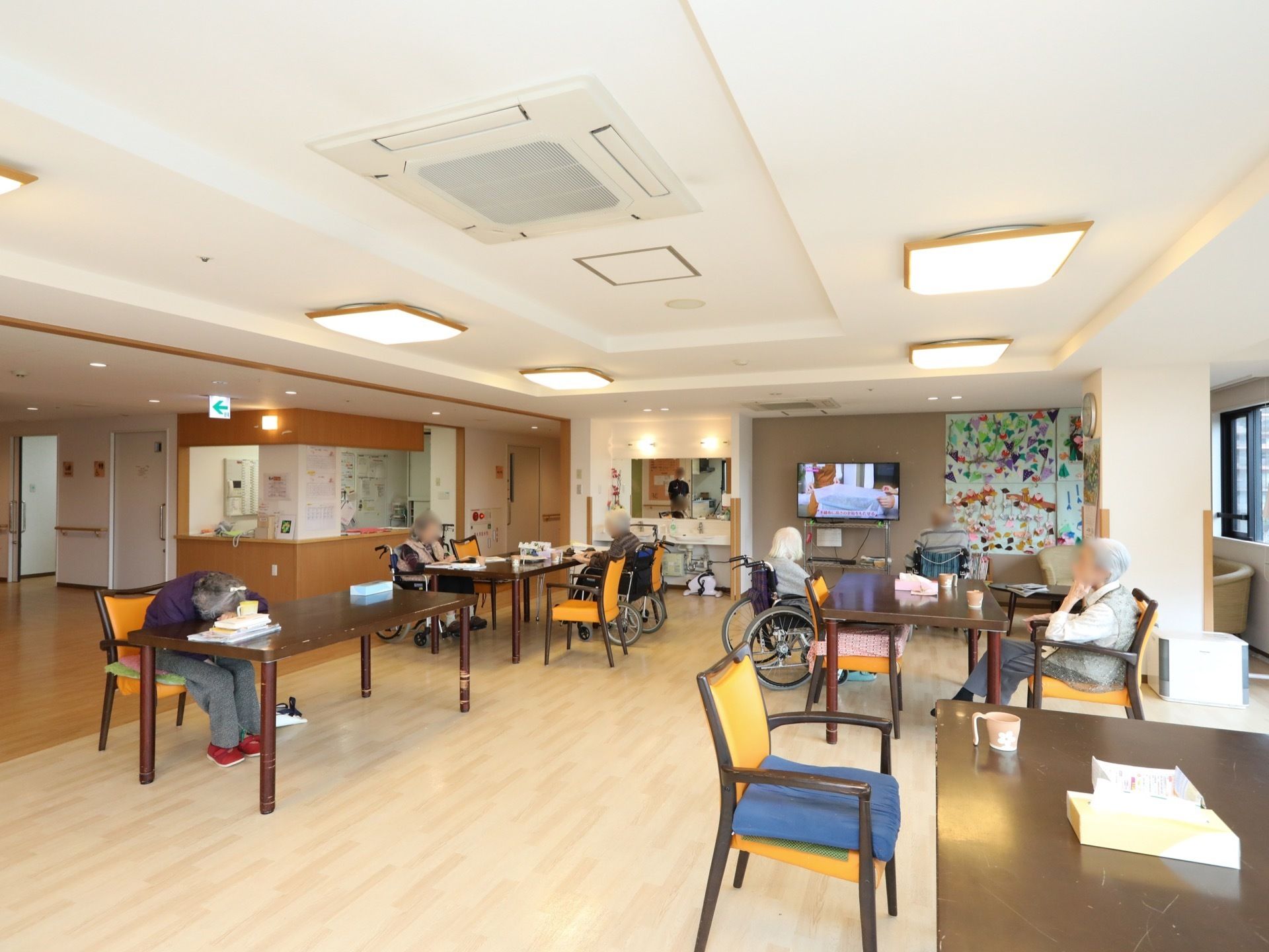 空調の付いた明るいホールがある。テーブルセットが並べられており、入居者様が席についてテレビを観ている。