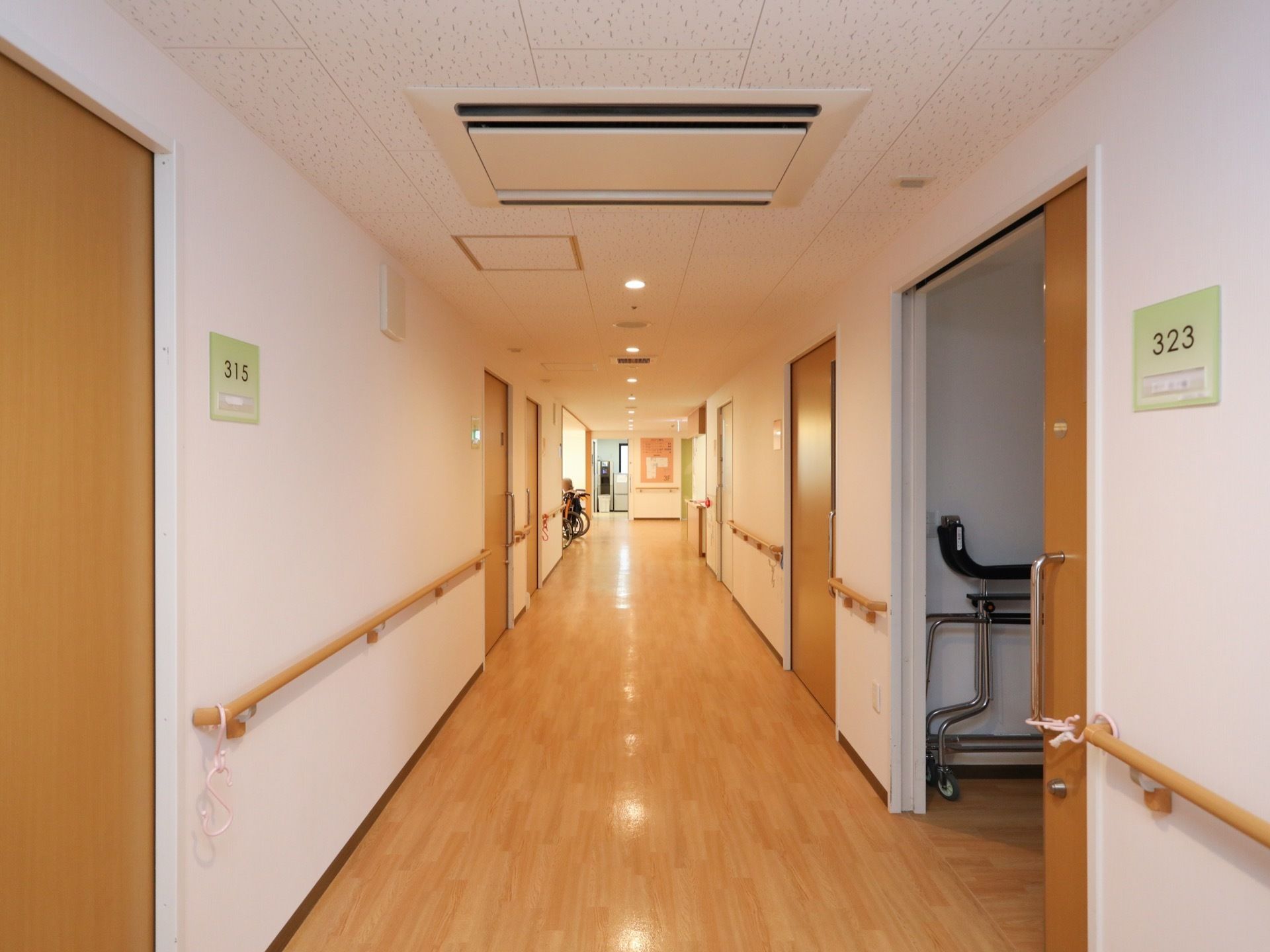 フローリングの廊下がまっすぐ続いている。天井には空調が付いており、壁には木目調の手すりが設置されている。