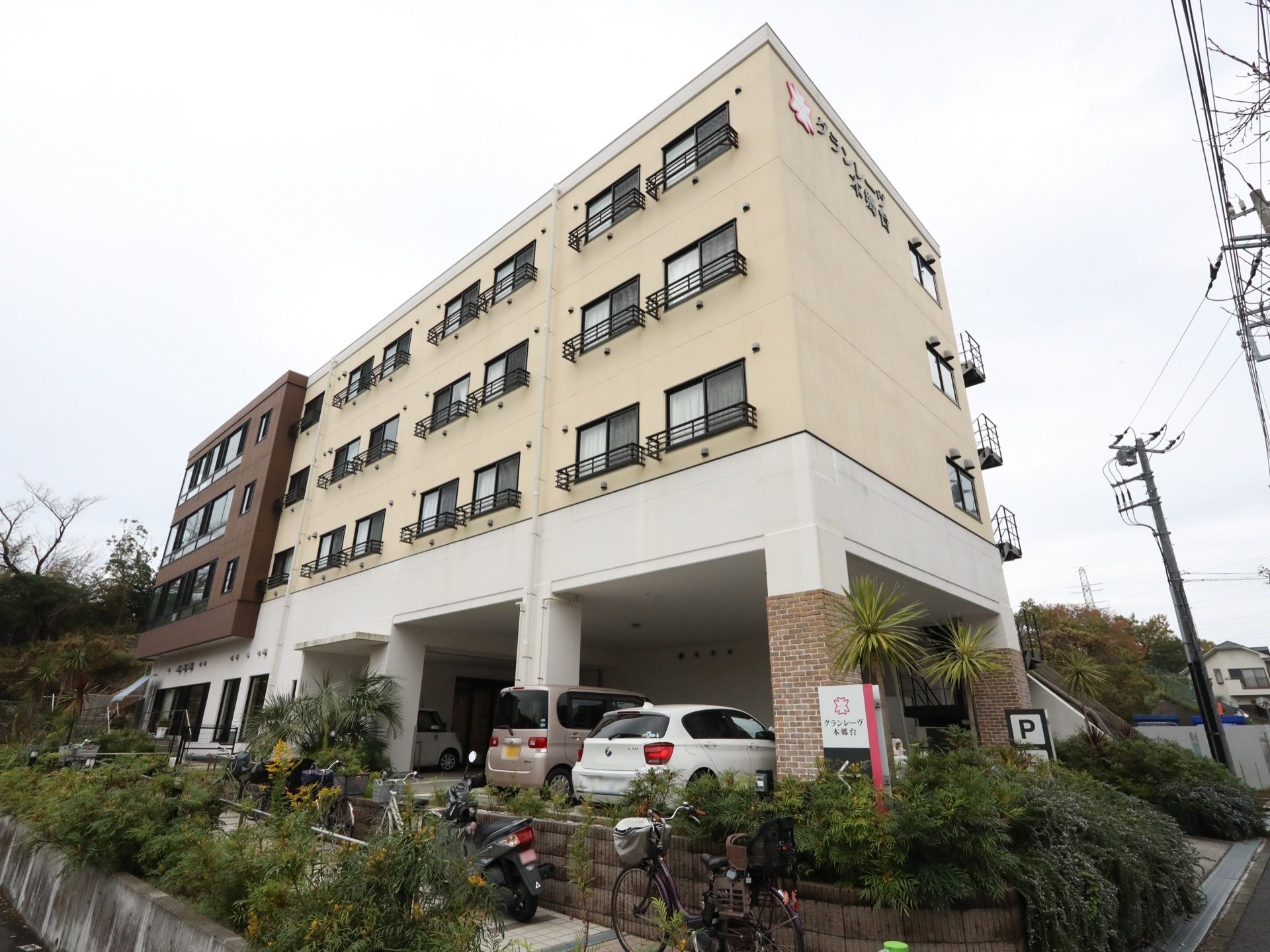 植栽に囲まれた４階建ての施設がある。１階部分は駐車スペースと玄関になっており、車が数台停められている。
