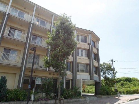 マンションのような４階建ての施設。手前に植え込みがあり、建物と同じくらいの高さの木が一本生えている。
