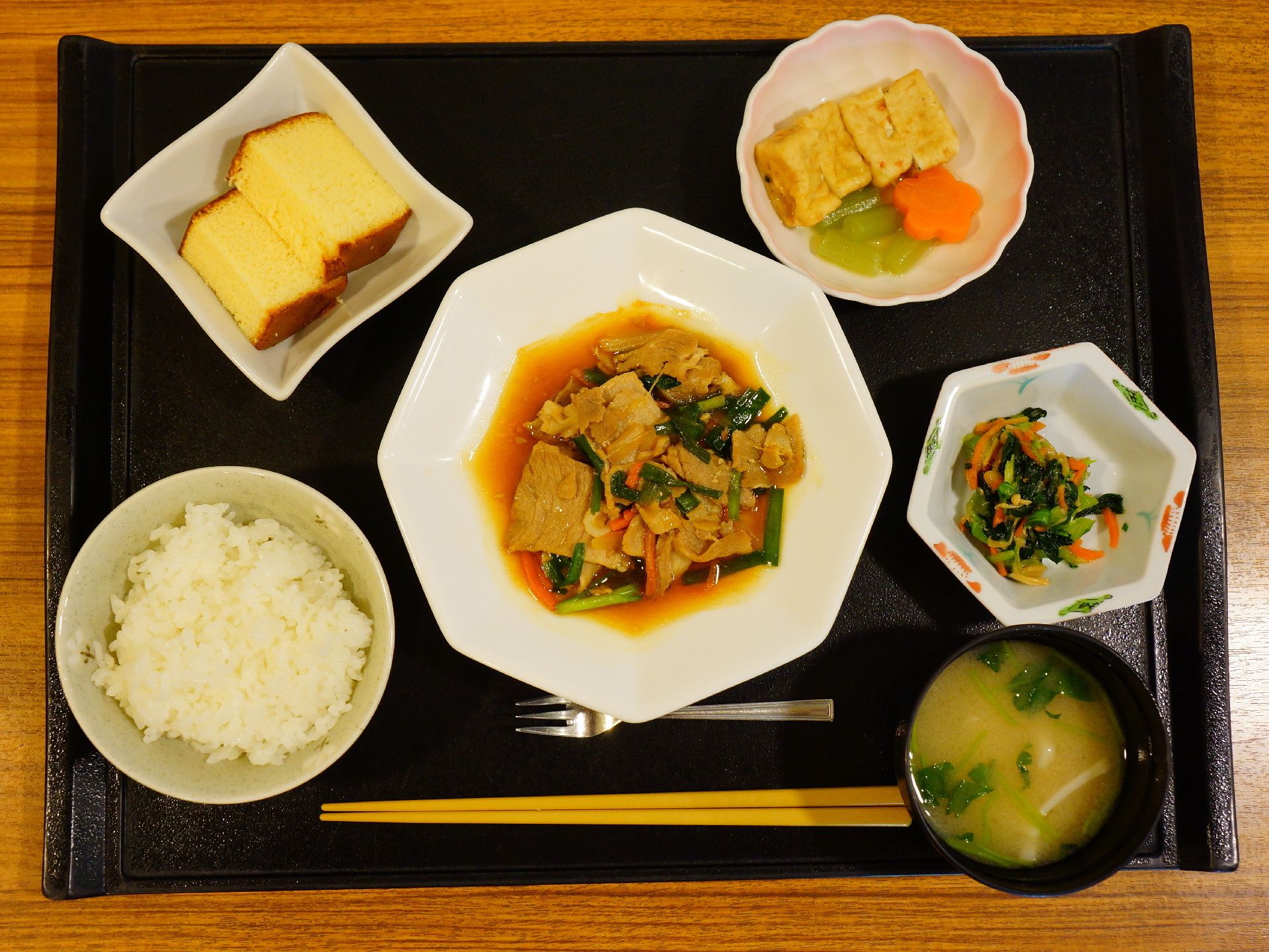 ごはんとみそ汁に、おかずが付いた食事。おかずは肉野菜炒めや、卵焼きがあり、デザートにバームクーヘンが付いている。