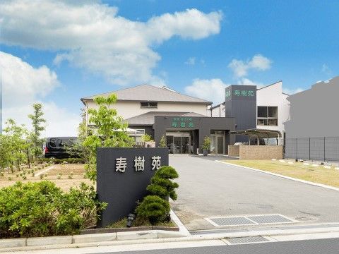 看板の周辺に緑が植えられていて、奥に施設の建物がある、敷地入口から見える風景