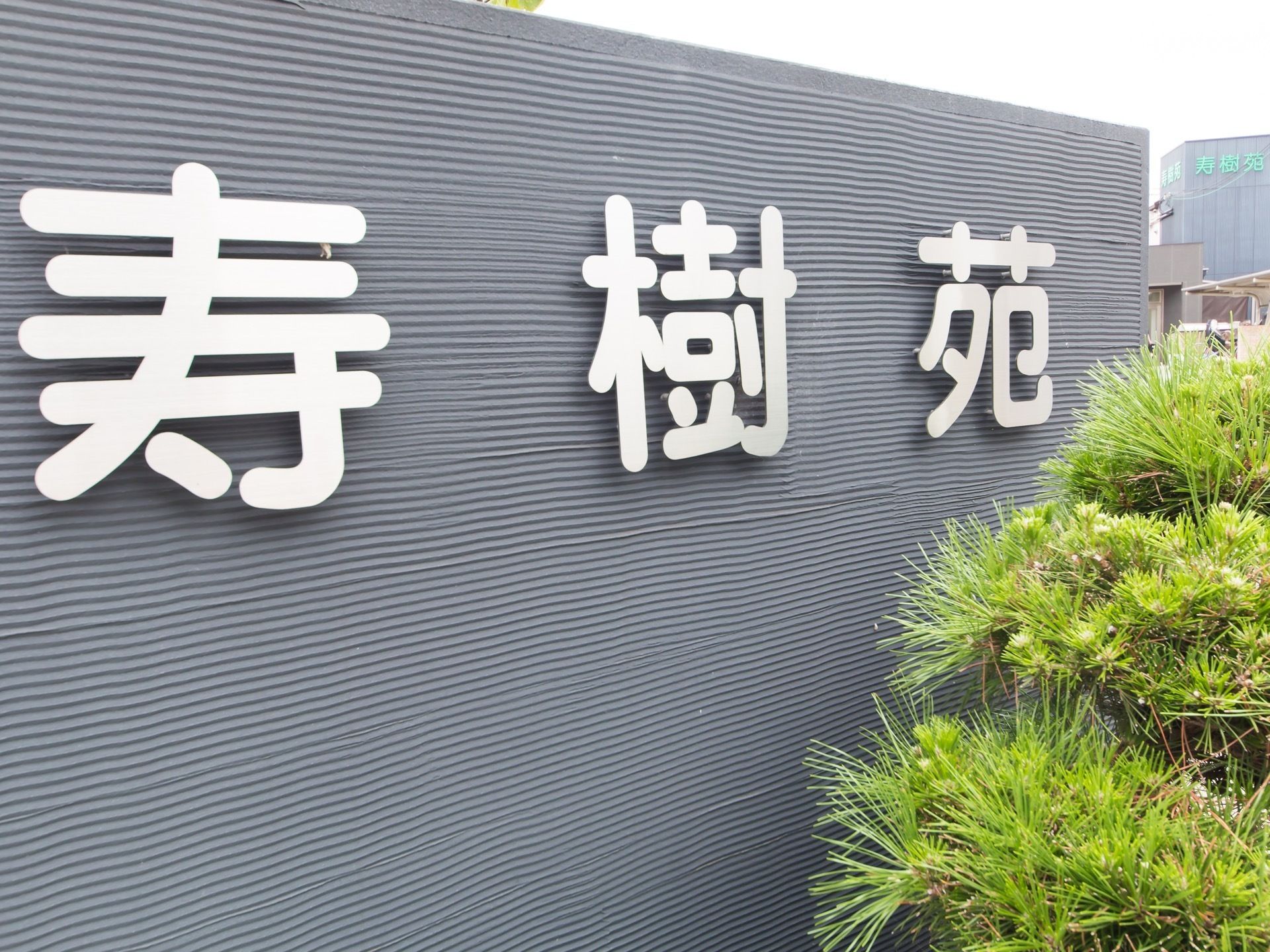 松の木の奥に黒い塀があり、一文字ずつ打ち抜かれた銀色の大きな施設名の文字が、等間隔に付けられている。