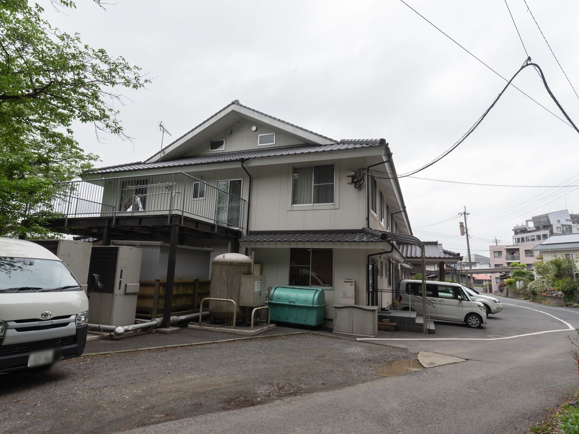 白い壁に瓦屋根の二階建ての建物である。出入口の階段には手すりが備えつけられ、施設前には車が駐車されている。