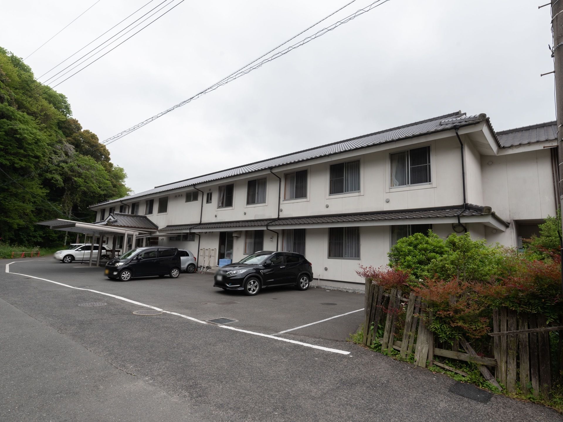 白色の壁に瓦屋根の２階建ての建物である。建物前には車が駐車できるスペースが確保されている。建物周囲は自然に囲まれ、静かな環境である。
