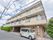 道路に面した鉄骨造3階建ての建物。薄いグレー色と茶系の外壁、建物の前には植木が植えられ、反対側には施設の車が止まっている。