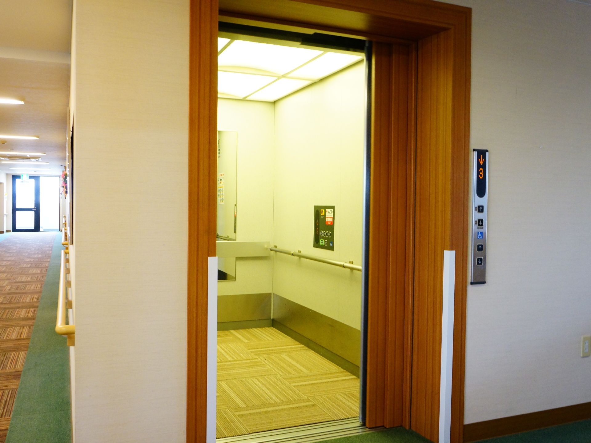 エレベーターは木目調の扉になっている。中は車いすで入っても十分な広さが確保されている。手すりも設置されている。