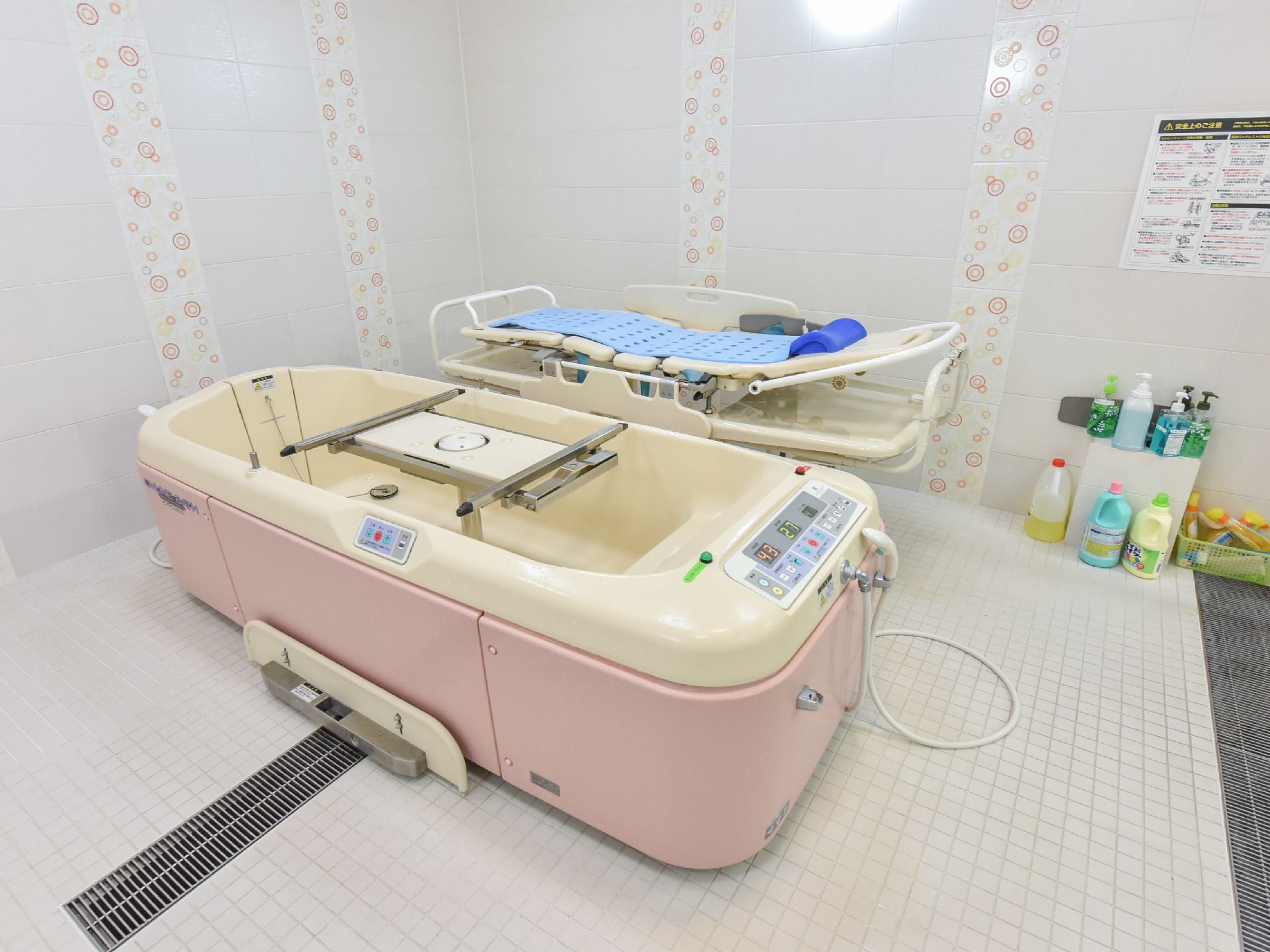 白色に縦帯の模様が施された壁。側面がピンク色の浴槽、その隣にストレッチャーが置いてある。端の壁際にはソープや洗剤が置かれている。