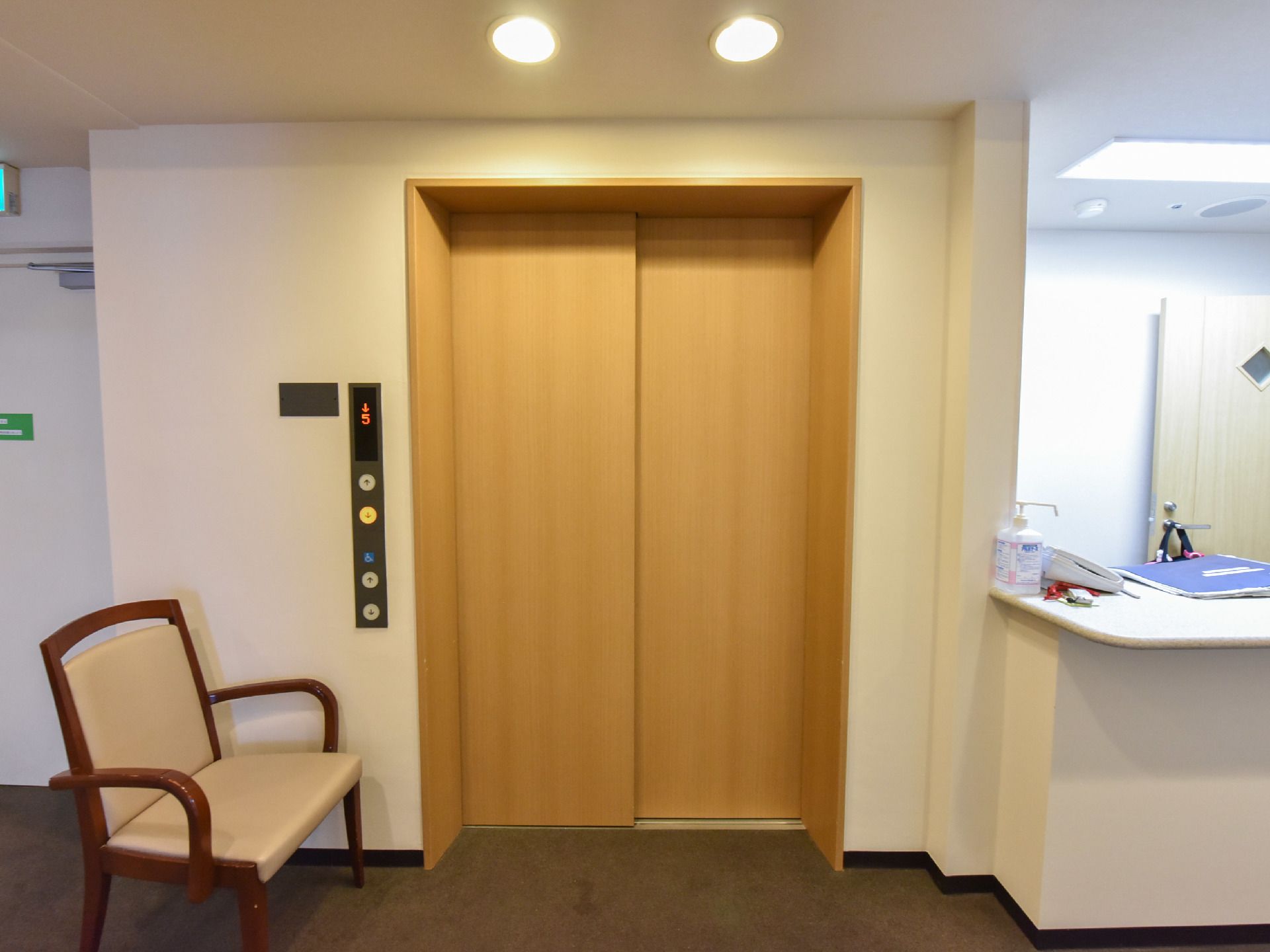 白色の天井と壁に黄土色系のエレベータードア。エレベーターのボタンの傍に一人掛けの椅子が置かれている。天井には、2つの照明を設置。