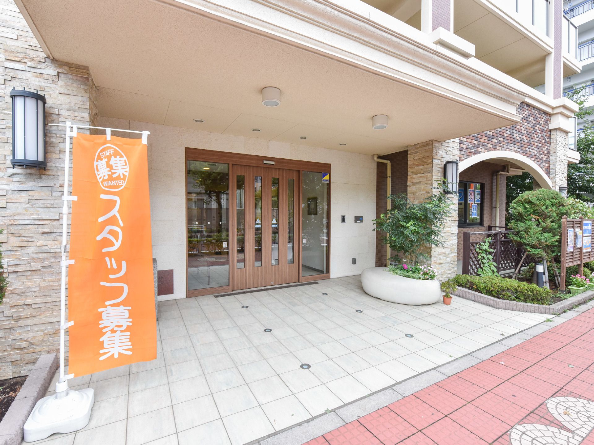 木目とガラス張り仕様の屋根付き玄関ドア。前方には植物が植えられ、道路傍にはスタッフ募集のオレンジの旗が立ててある。