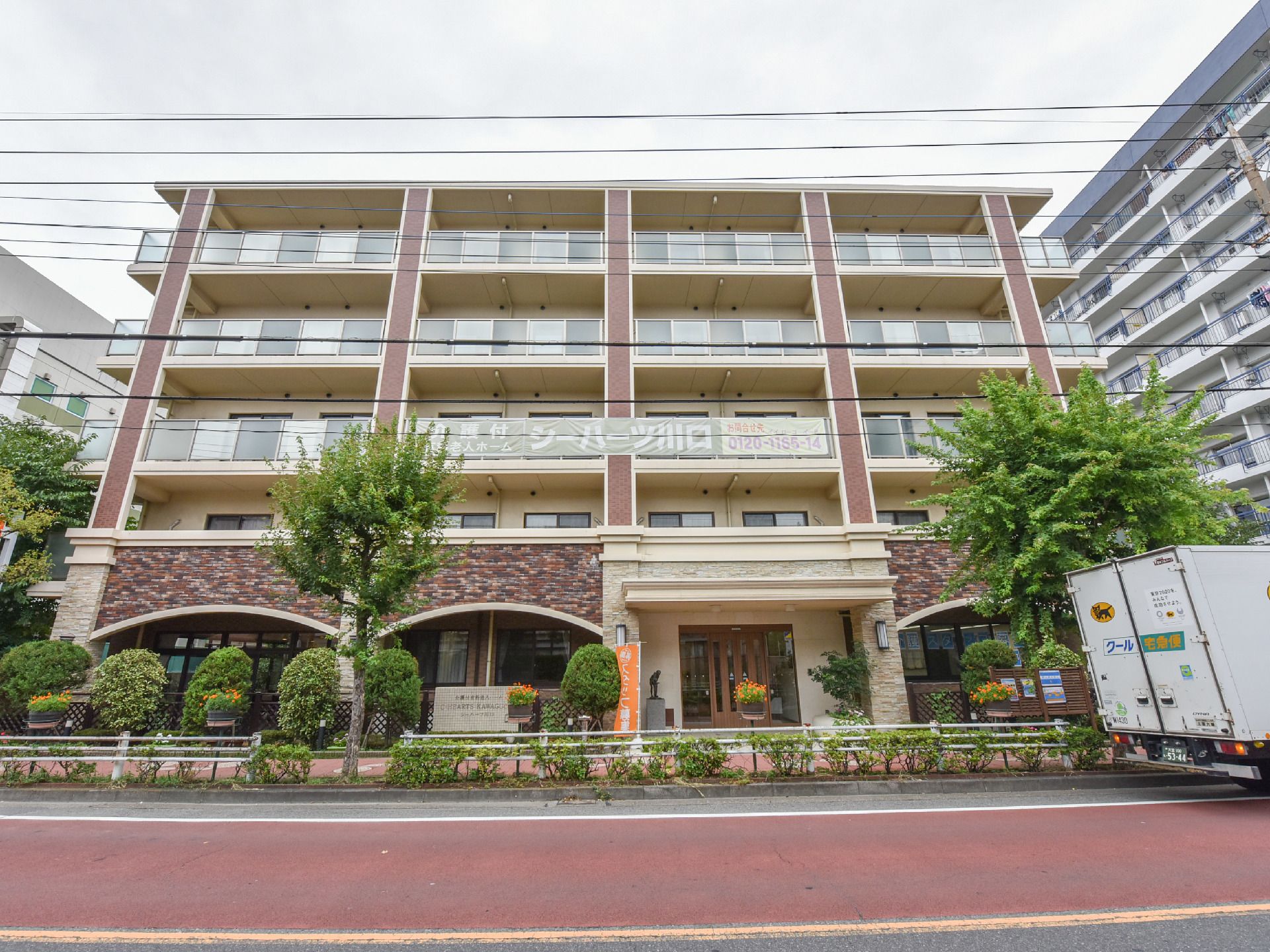 ベージュ色の外壁に1階のデザイン仕様の設計の外観。ホテルの様なエントランスの5階建ての建物。建物前は、歩道を挟み大通りとなっている。