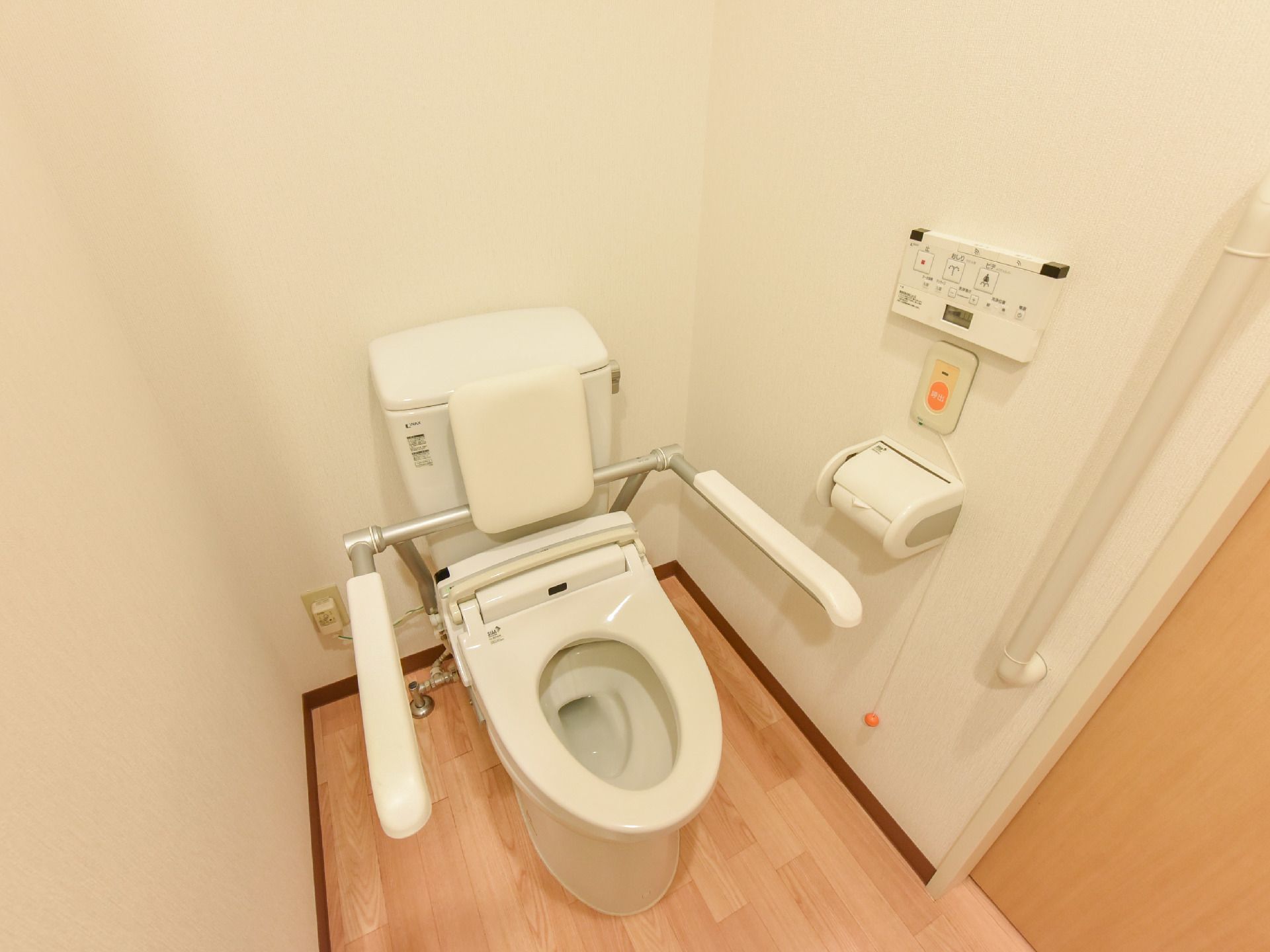 白壁に囲まれた便器。両側には手すりが設置してあり、出入口の傍には、縦長の手すりが付いている。便器横の壁にはナースコールが設置。