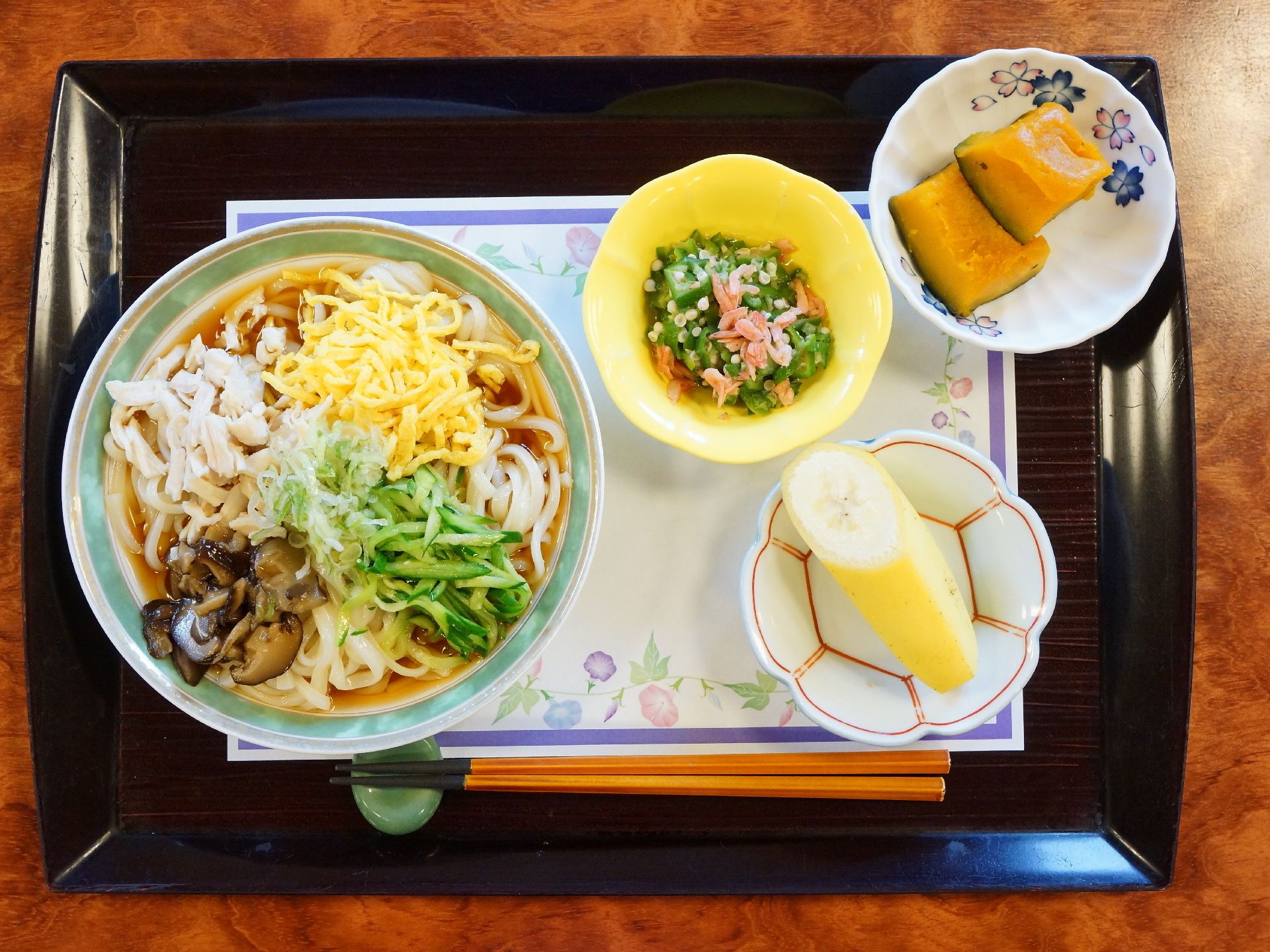 トレーの上に具材が盛り沢山の麺類、小鉢が2つとフルーツが添えてある。1日の栄養バランスを考慮された食事。