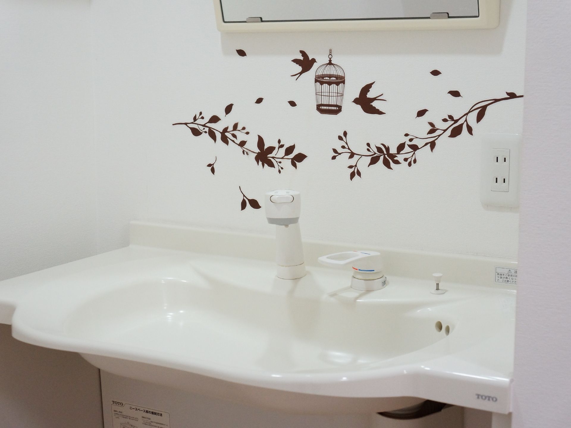 洗面台の鏡の下の壁には鳥かごに飛んでいく鳥や植物の絵の壁紙が貼られている。シャワーヘッドもついているので便利。