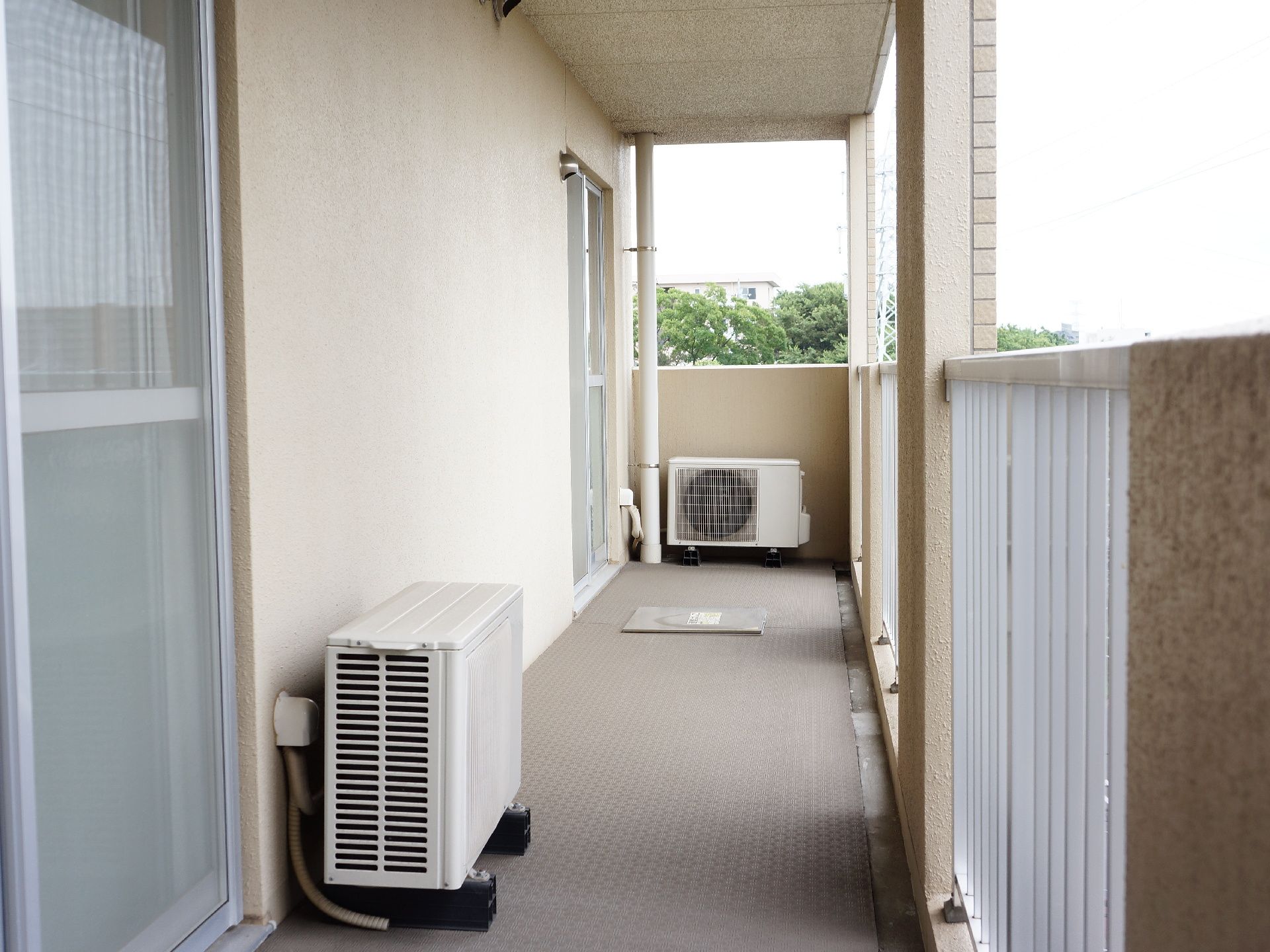 ベランダにはエアコンの室外機が置かれている。床には避難用の入口がある。十分な高さの白い手すりがついている。