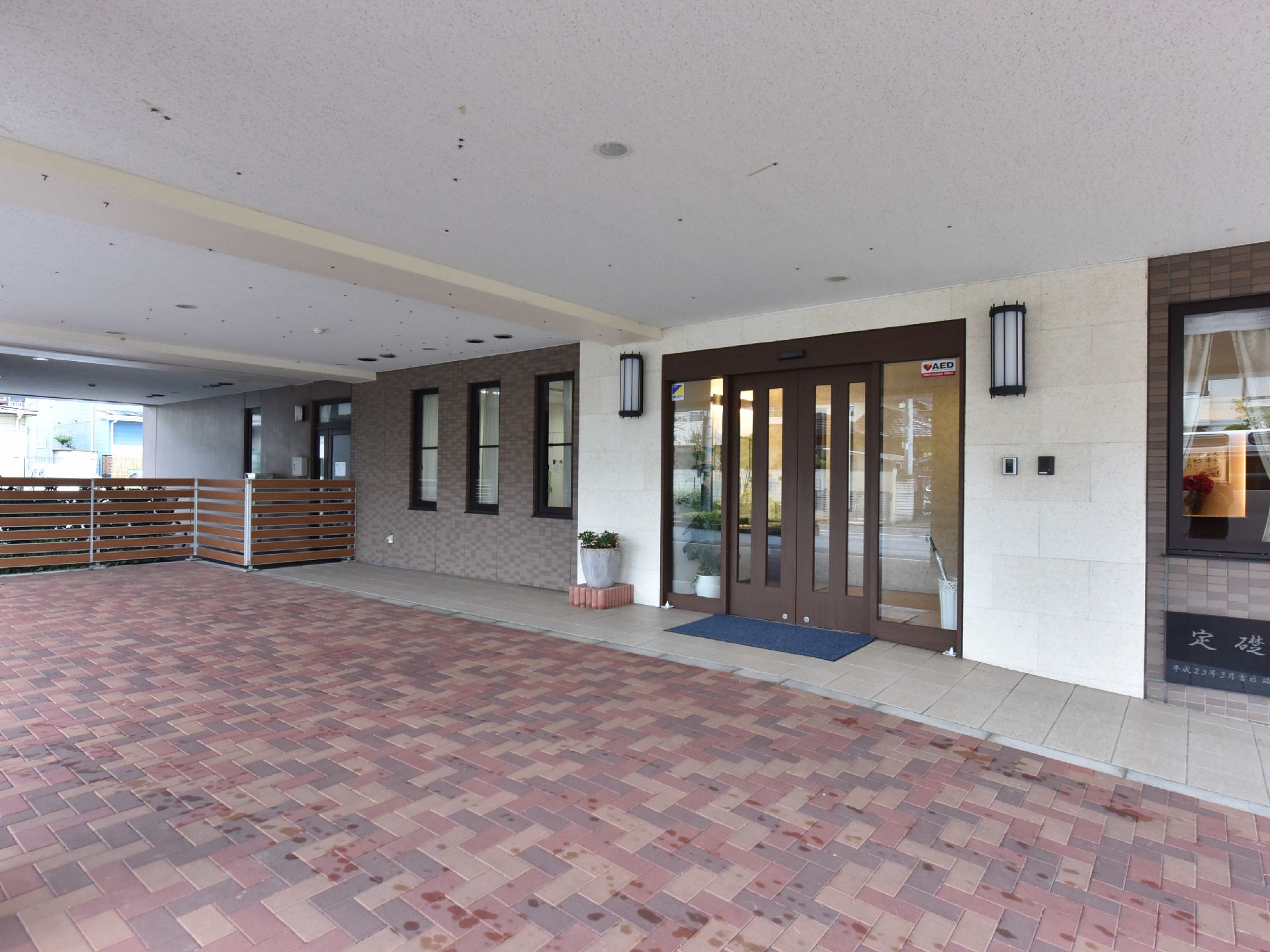 施設入口は大きさと色が異なるタイルの床になっている。茶色い扉の横側の外壁には定礎と書かれた石がはめ込まれている。