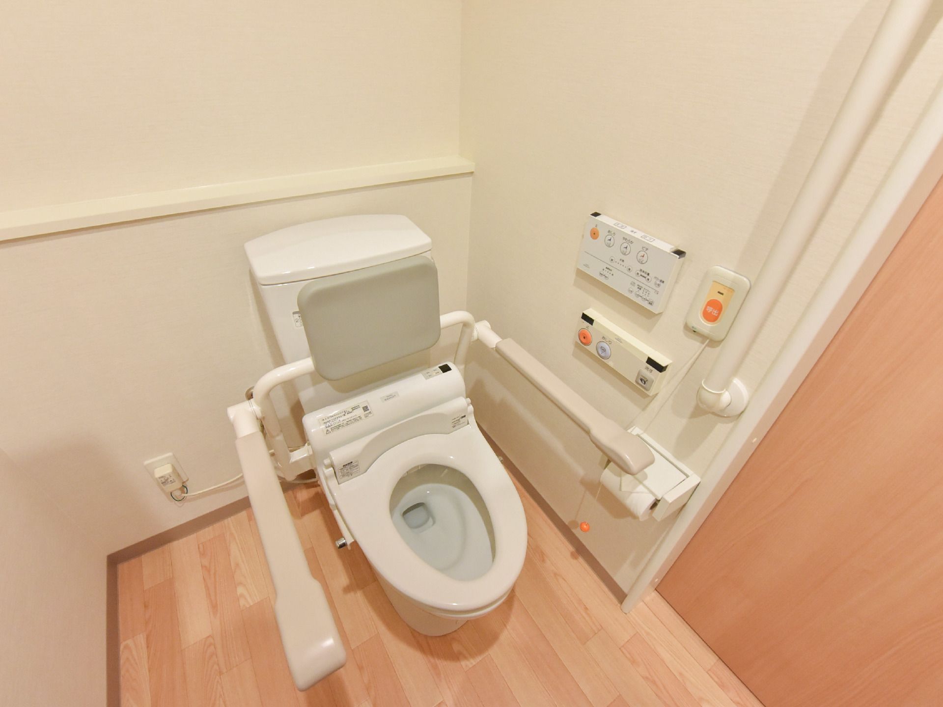 トイレの便器の両サイドには肘置きがついている。肘置きと同じ素材の背もたれも付いている。壁側には呼び出しボタンが設置されている。