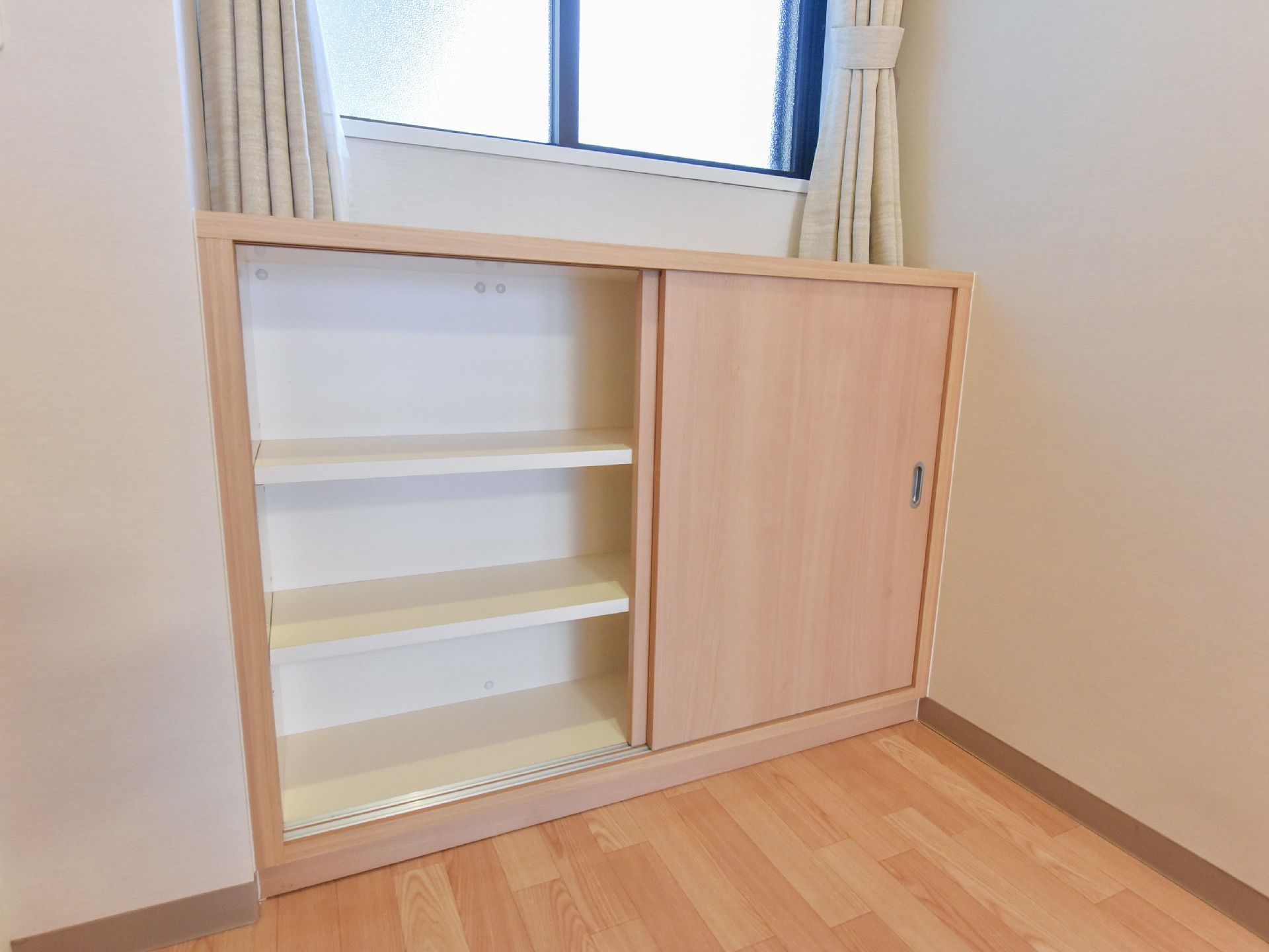 結ばれたカーテンがかかった窓際には茶色い収納棚がある。収納棚は窓枠とほぼ同じ幅で、中を開けると3段に仕切られている。