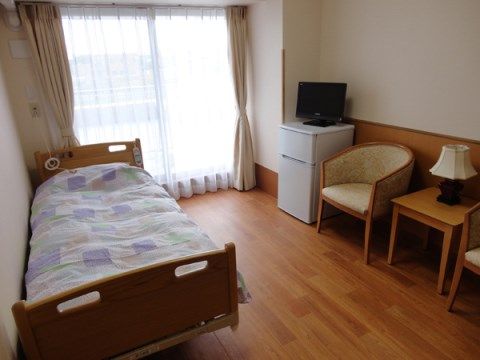 フローリングの室内に、ベッドやテーブル、椅子、冷蔵庫、テレビが置かれている。ベランダへ出られる大きな窓がある。