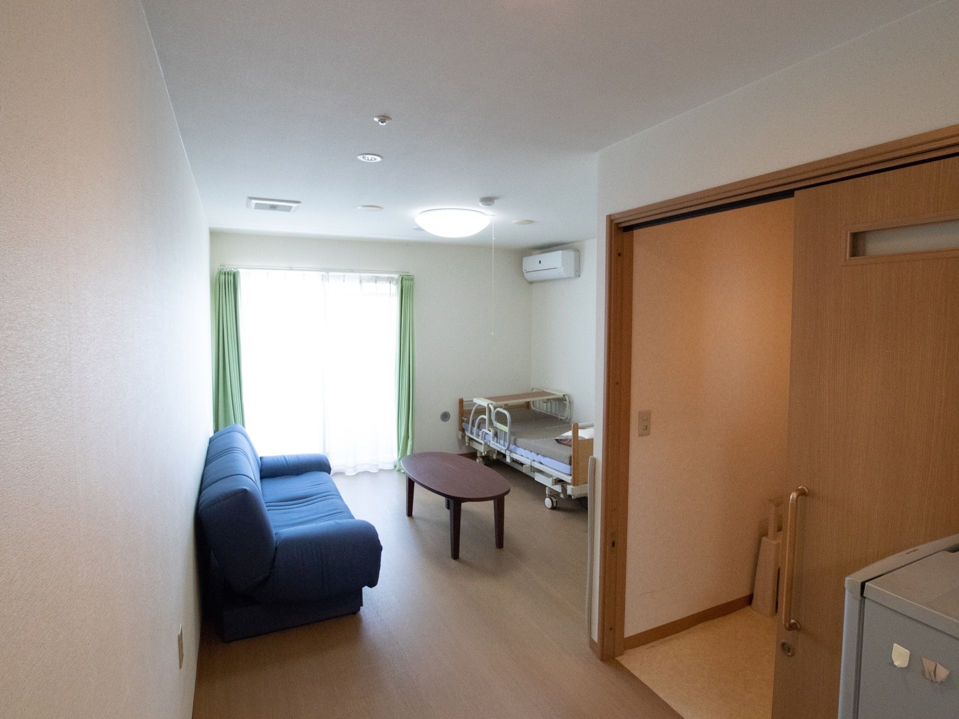 右手には引き戸のトイレがある。奥には青色のソファと、茶色の低い机と布団の敷かれていないベッドが置いてある。空調設備があり、大きな窓もある。