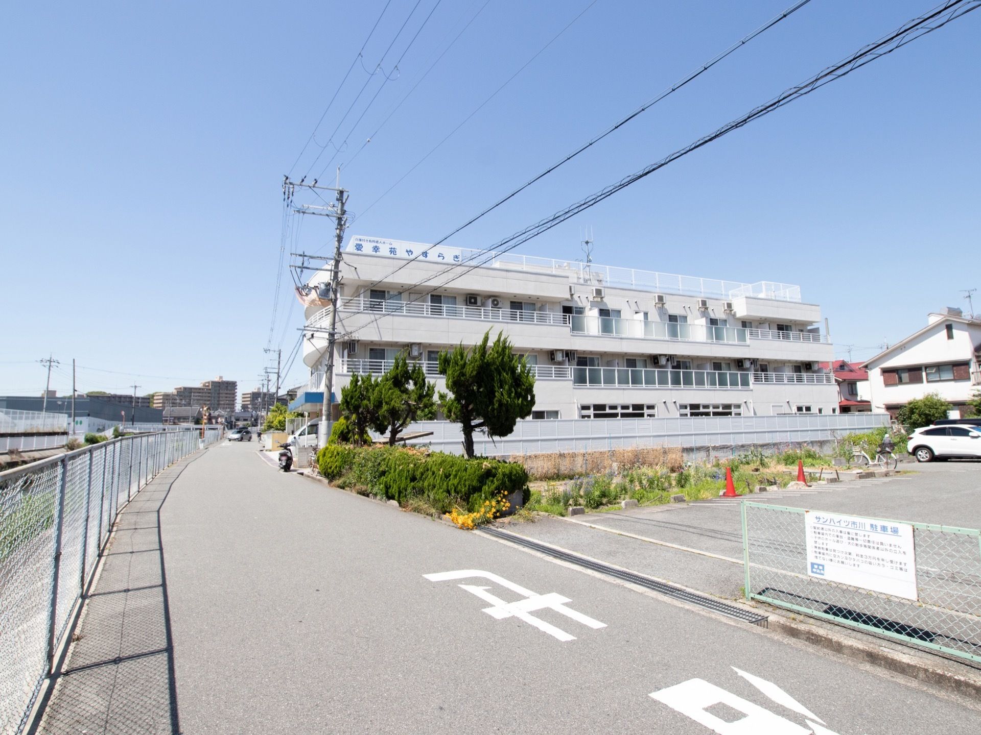 大きな三階建ての白い建物。屋上がついており、フェンスには施設名が書かれた看板がある。また、建物は広い道路に面している。