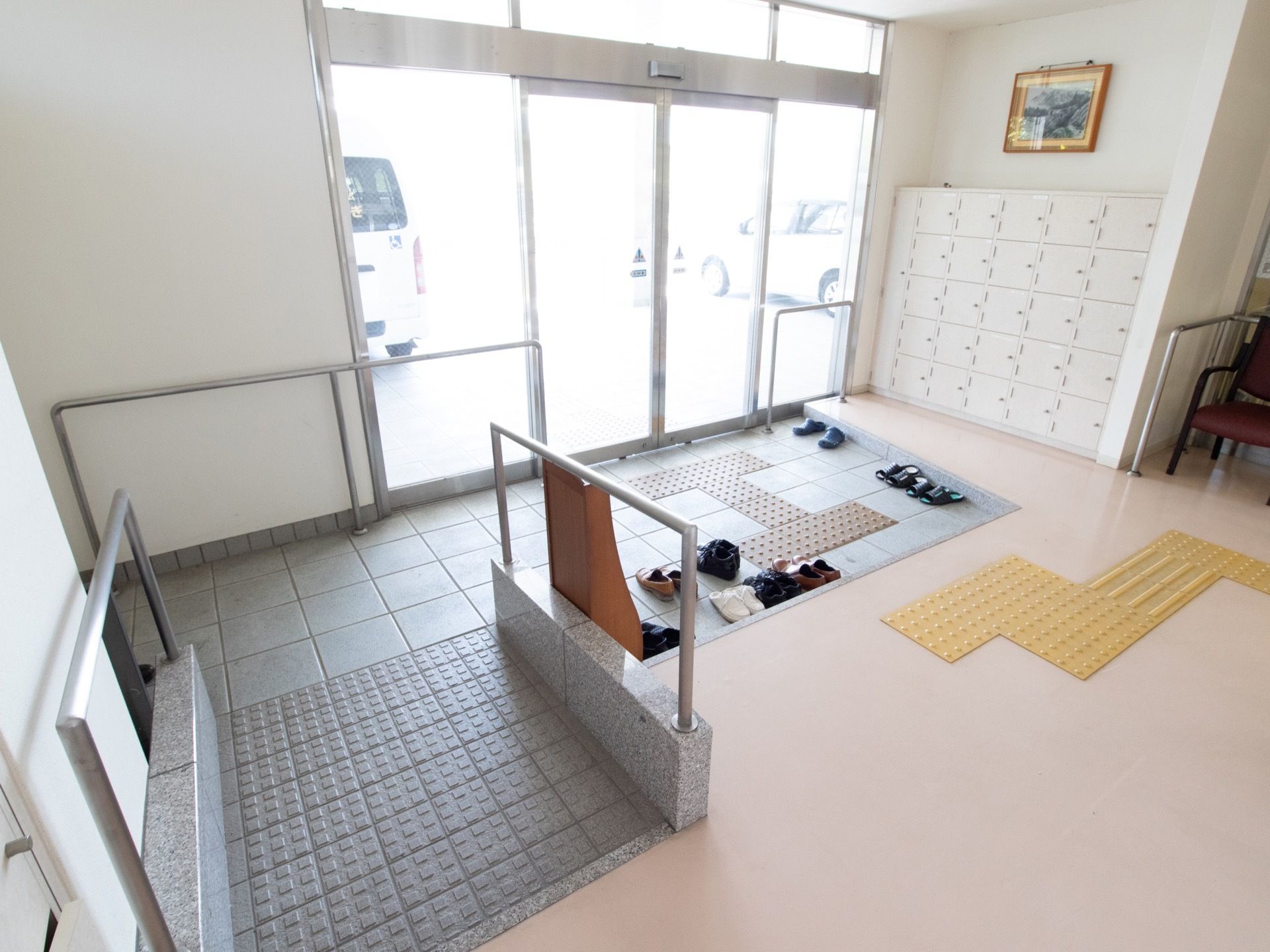 自動ドアがあり、右手には大きな靴箱が見える。玄関内には点字ブロックが敷かれており、左手には手すり付きスロープがある。