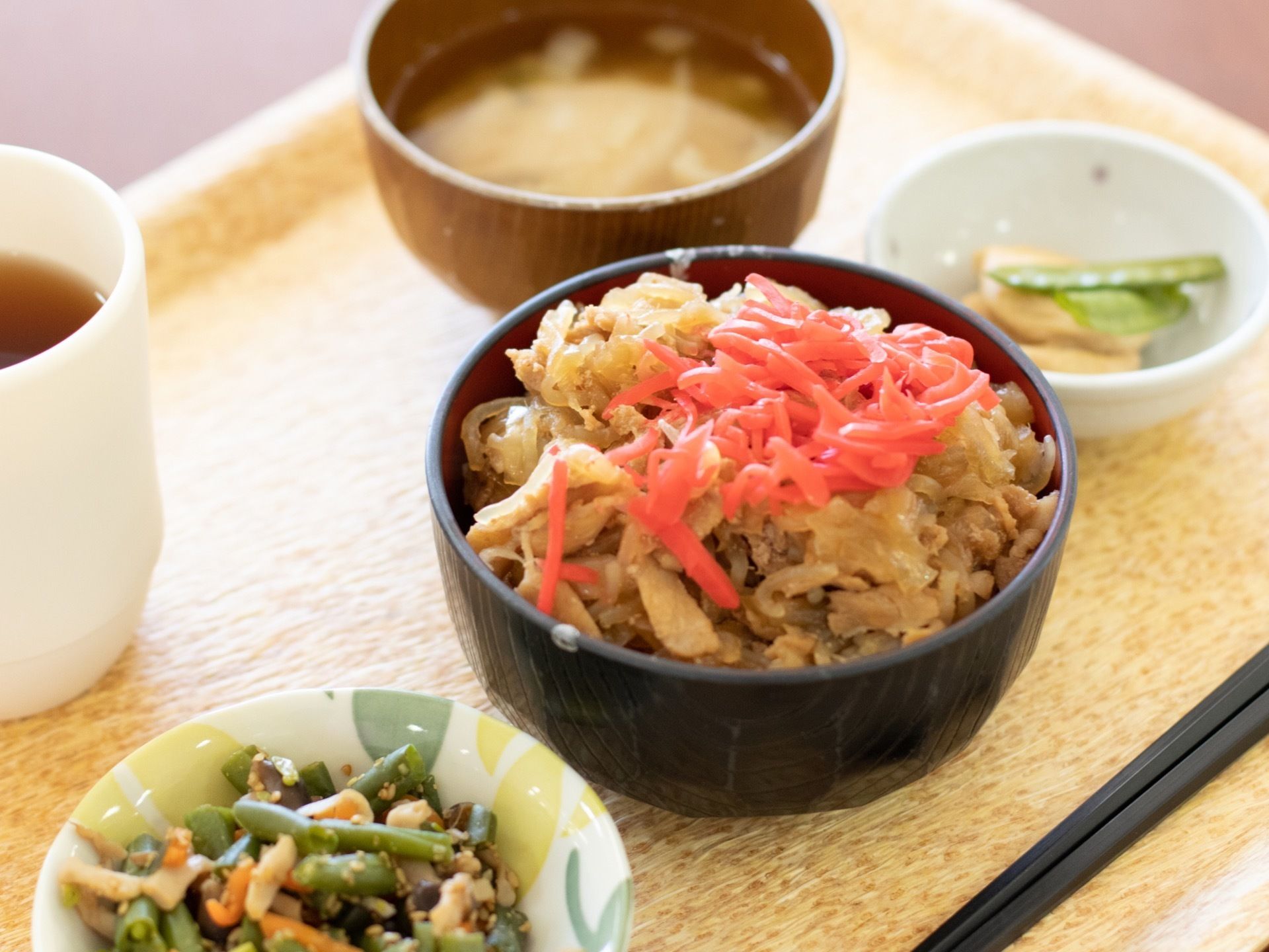 カップに入ったお茶、木の器に入った味噌汁、小鉢に入った野菜類二種、器に盛られた牛丼、黒い箸が一膳木のトレーにのっている。