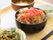 黒い器に紅生姜ののった牛丼が盛られている。また、野菜系の小鉢が二種あり、手前には黒い箸が用意されている。