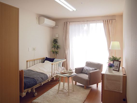 フローリングの居室には電動ベッドやタンス、エアコンが完備されている。ベランダへ出られる大きな窓にはカーテンが付いている。