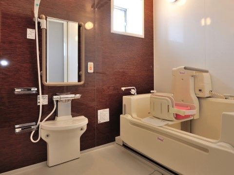 壁には鏡が付いており、そのまわりには手すりが備え付けられている。介護浴槽にはピンクのクッションが付いた椅子が設置されている。