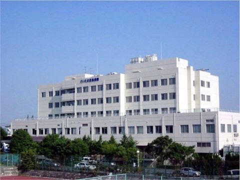 白い大きな建物で窓がたくさんあり、上のほうに青い字で病院名が書かれている。道路に沿って木が植えられており、車が所々に停まっている。