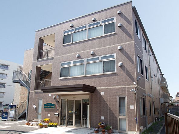 玄関周りに花の咲いた鉢植えが並ぶ、3階建てで奥行きのある施設