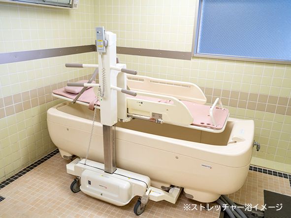 広い浴室に白い介護浴槽とストレッチャーが設置されている。壁の上のほうには窓があり、天井には換気扇や照明が付いている。