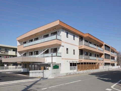 オレンジ色と白色の明るい外観の３階建ての建物が建っている。敷地はアスファルトで舗装され、コンクリートの壁に手すりが設置されている。
