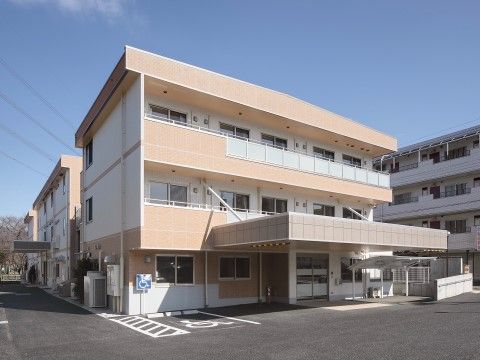 ３階建ての建物の前に屋根の付いた自転車置き場がある。舗装された敷地に白線で障害者用駐車スペースの表示がされている。