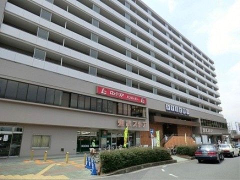 白いショッピングモールのビルが建っている。数本の黄色いのぼりが立てられ、建物の前にタクシーが２台停められている。