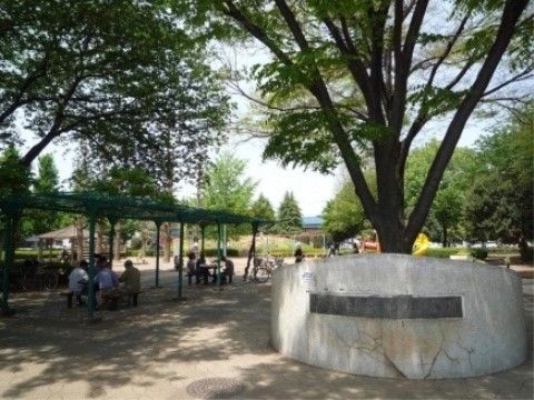 大きな樹木がたくさん植えられた公園に木陰ができている。数名の人々が、屋根のあるベンチに腰かけている。