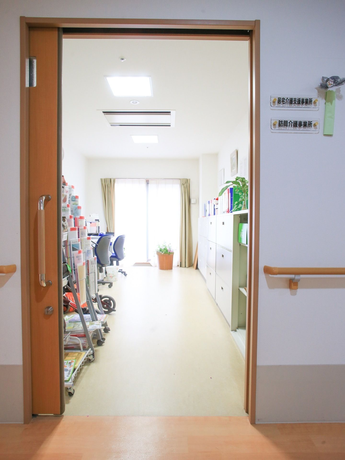 事務机と椅子と書類棚が置かれている。各種のパンフレット類がある。観葉植物が飾られている。窓にはレースと黄土色のカーテンが掛けられている。
