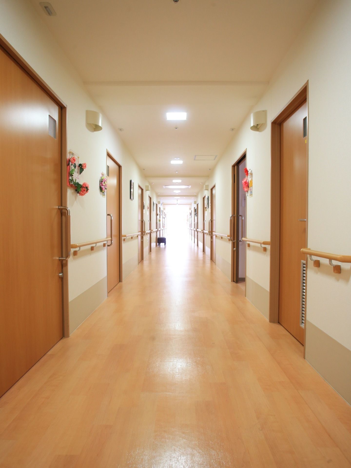 壁には手すりが取り付けられている。居室のドアは引き戸式である。部屋の前には造花などが飾られている。通路奥には窓がある。